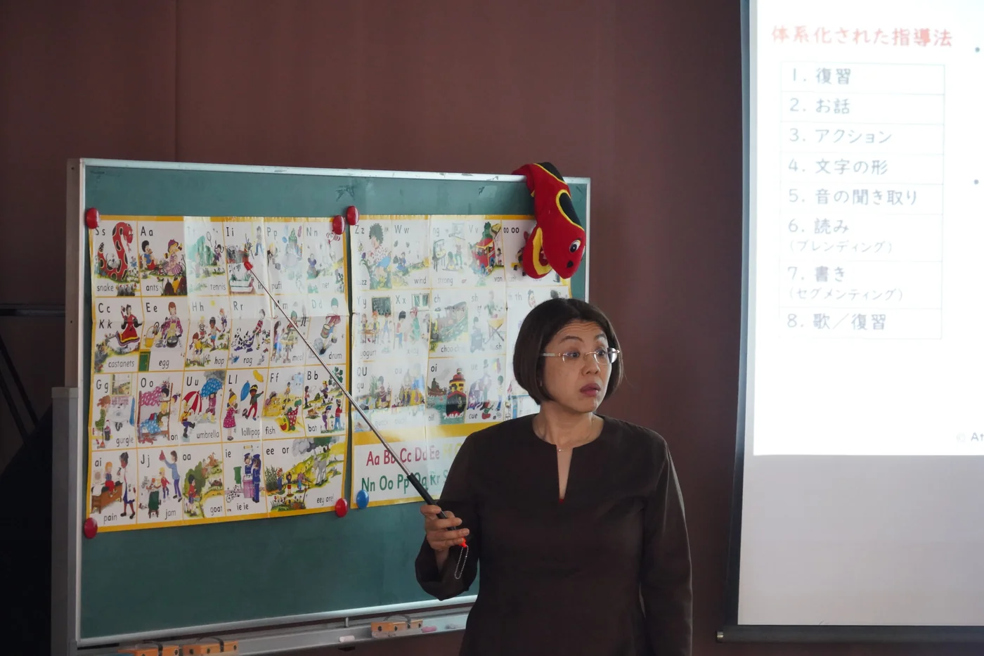 Atsuko Maruyama explaining with a phonics chart and teaching-sequence slide at Moriyama Library
